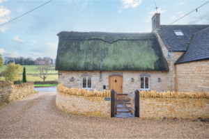 The front of the Studio cottage with Cotswold stone walls a thatched roof and a strong wood front door and gate with a background of a sunny day with a green field.
