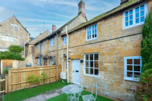 The back garden of the Honeypot Cottage with Cotswold stone walls and well-maintained grass in front. A traditional outdoor dining table in a white finish is on the grass to the right of the path.