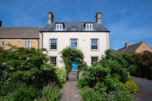 The front of Kissing Gate Cottage that is a tall white wash building with well-kept bushes in the foreground and framing the front door.